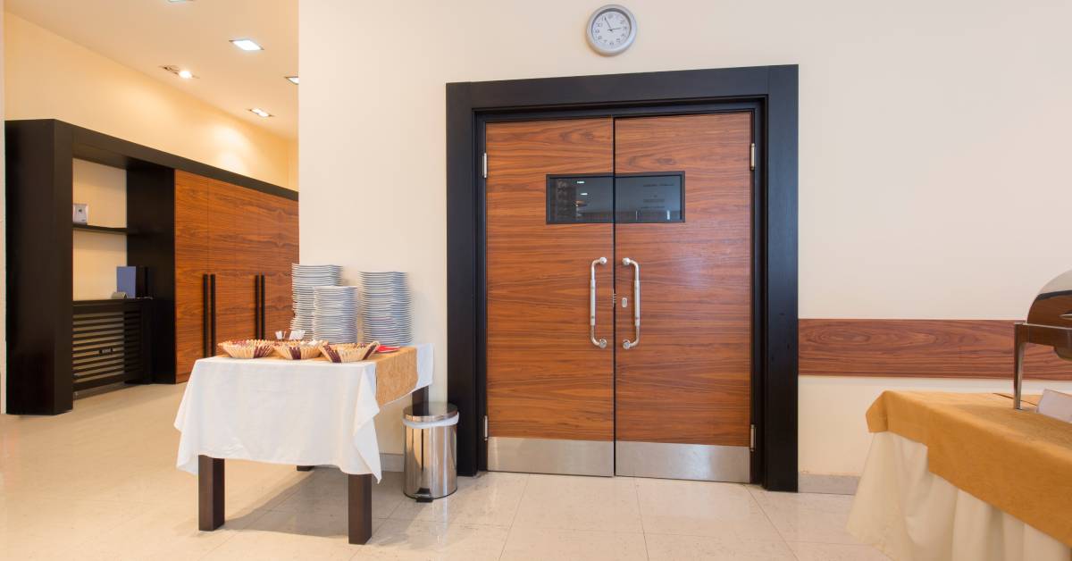 A brown swinging door with handles. There is a table full of neatly folded napkins next to the pair of doors.