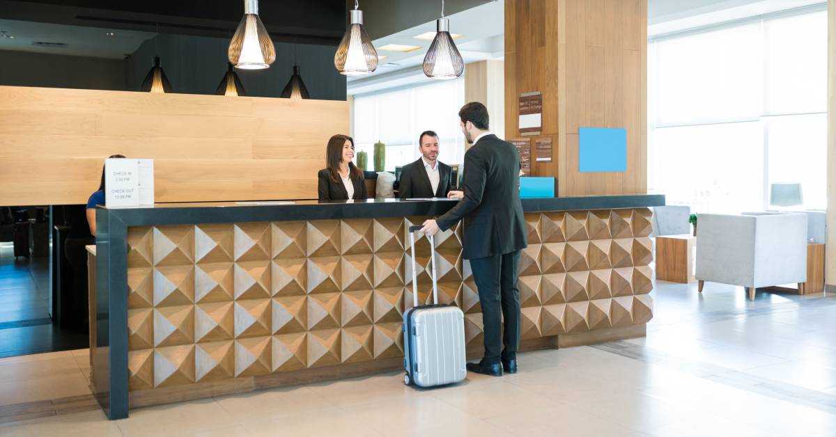 Two clerks working at a hotel reception desk. There is a man in a suit talking to the two clerks at the counter.